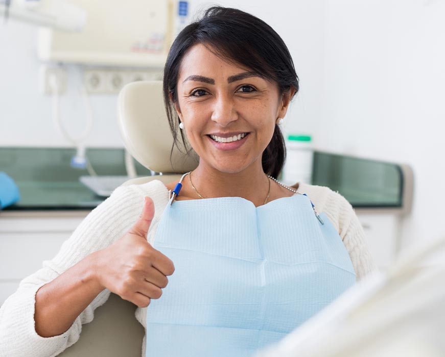 A woman, presumably a dental hygienist, sitting in front of a dental chair, smiling and giving a thumbs-up gesture.
