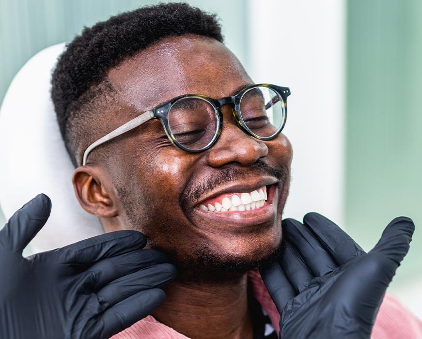 Man with glasses sitting in dental chair, smiling broadly, with hands clasped together over mouth.