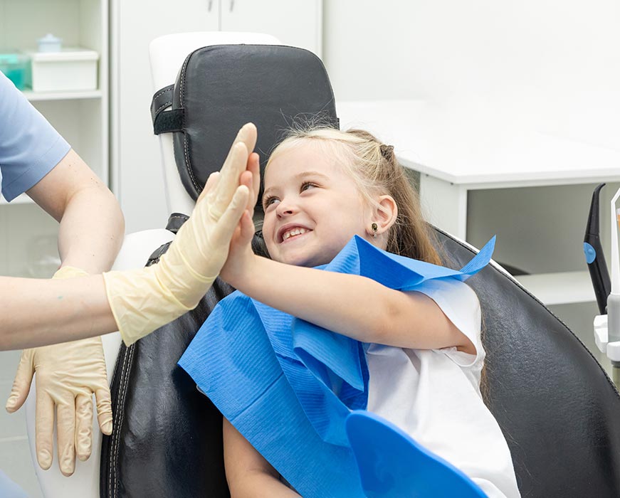 A young girl sitting in a dental chair, smiling at the camera while a dental professional gives her a thumbs up.