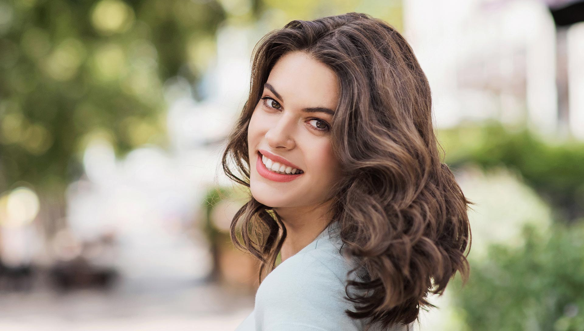 The image features a smiling woman with dark hair wearing glasses, seated in a dental chair with a dental professional s hand holding a dental instrument near her mouth.