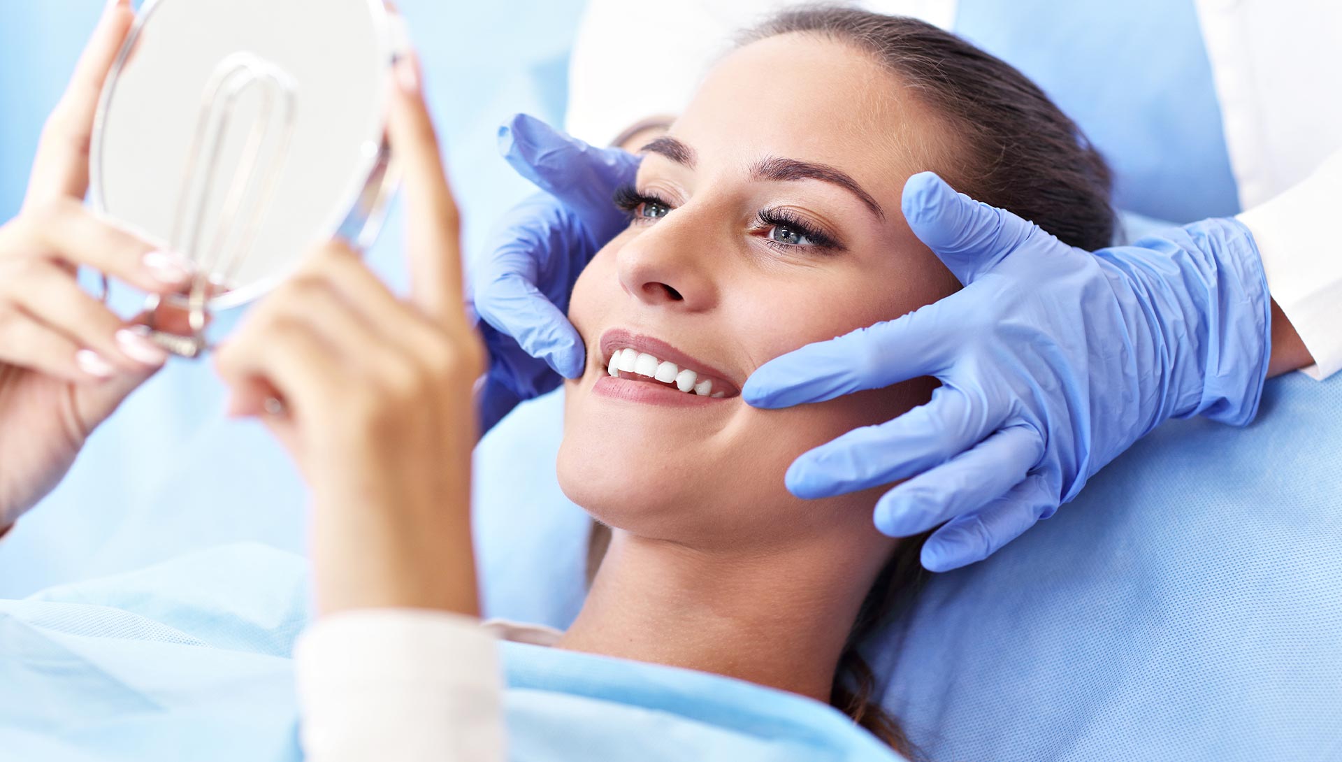 A woman in a medical setting is receiving a cosmetic procedure with a smiling expression while holding up her hand.