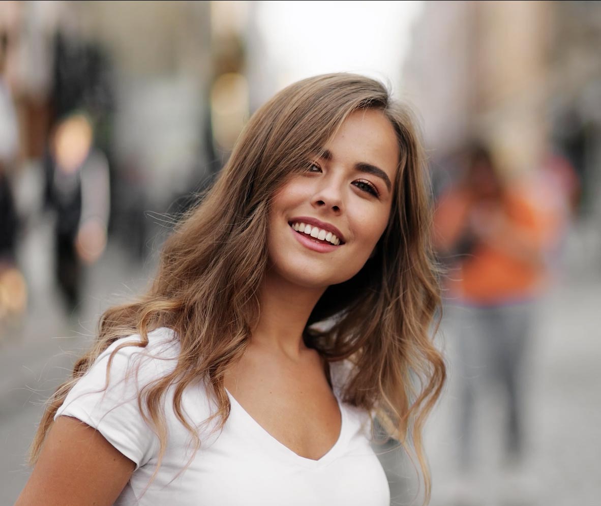 The image shows a young woman with long hair smiling at the camera while standing outdoors during daylight.