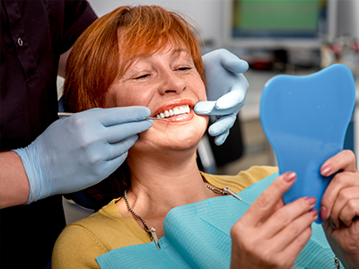 A woman seated in a dental chair with a blue tooth impression mold on her chin, smiling at the camera while looking at her teeth, with a dental professional attending to her.