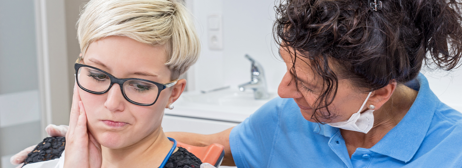 A woman with glasses, wearing a black shirt, sits in front of a dental hygienist who is holding a mirror over her face, both in a dental office setting.