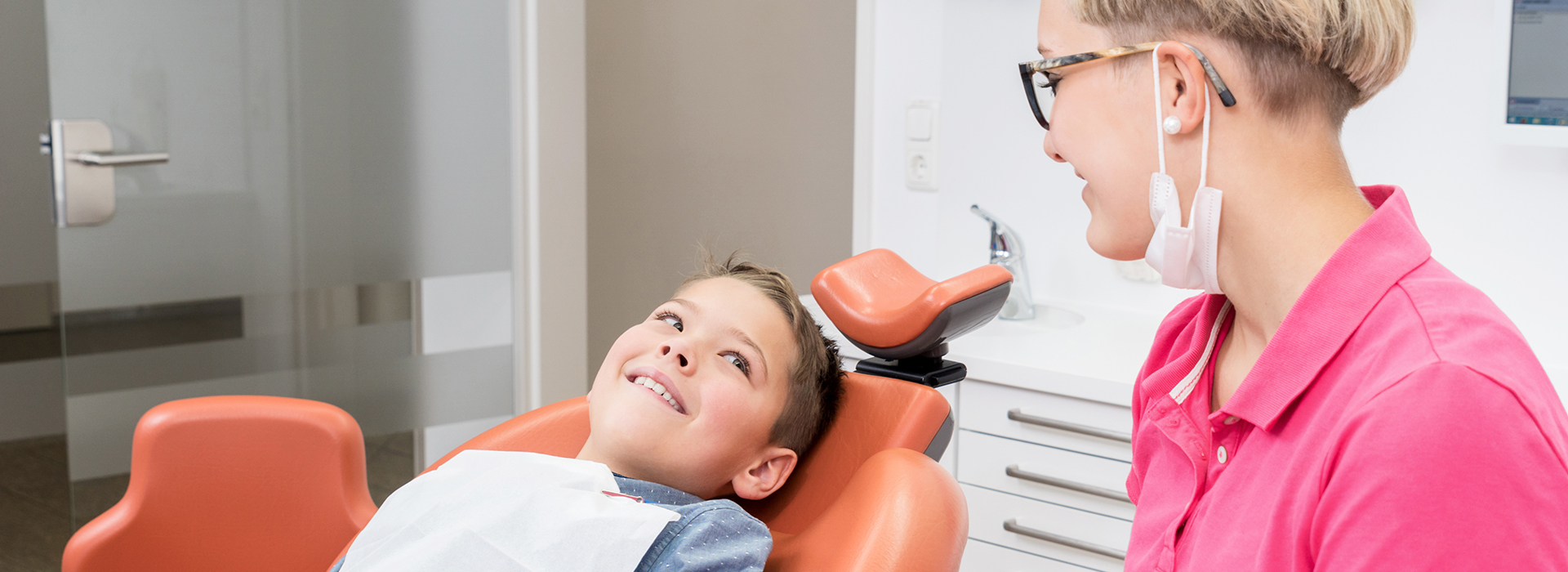 The image shows three individuals in a dental office setting  a woman seated in a dental chair with her mouth open, a young boy standing next to her smiling at the camera, and a woman standing behind them both, likely a dentist or dental hygienist, wearing a pink shirt.