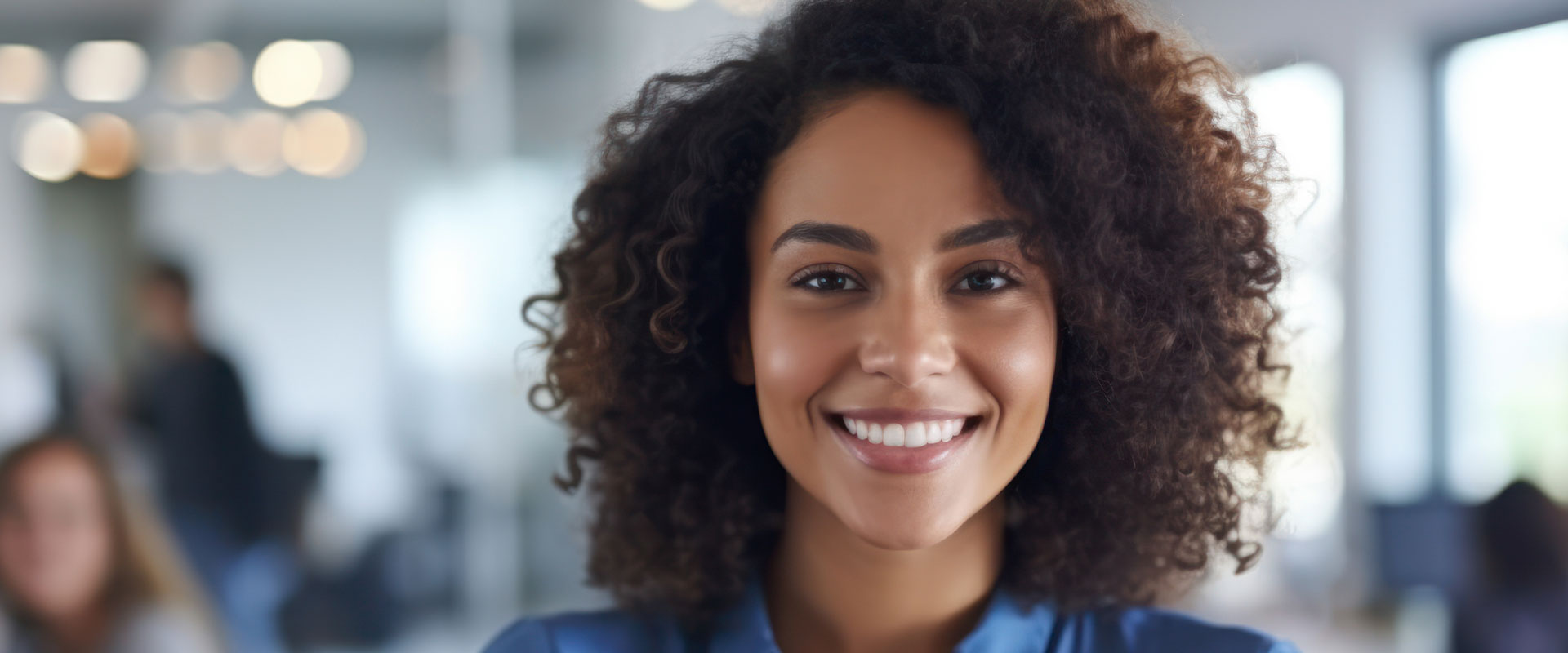 The image shows a person with curly hair smiling at the camera, wearing a blue top, against a blurred background of an office environment.
