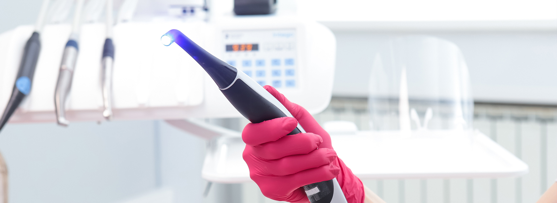 The image shows a person s hand holding an electronic device with a blue light, positioned in front of a white background with various medical equipment and a red glove.