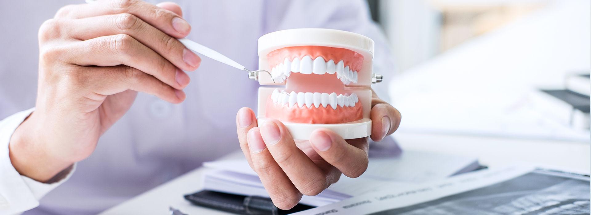 The image depicts a person's hand holding a toothbrush over an open mouth model, with another person examining dental implants on a table.