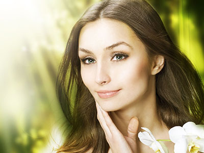The image features a woman with fair skin, wearing makeup, looking directly at the camera with a slight smile, set against a blurred background with a plant-like texture and soft lighting that highlights her facial features.