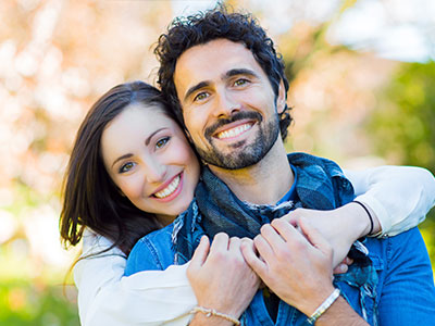 A man and woman are hugging each other outdoors  they both appear happy and are looking at the camera.