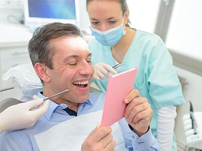 The image shows a man sitting in a dentist s chair with a cardboard sign on his face reading  Welcome to Dentistry  while a dental professional looks on, smiling.