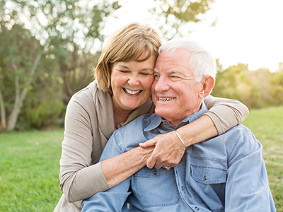 This is a color photograph featuring an elderly couple sharing a warm embrace. The man is wearing a blue shirt and the woman is in a light-colored top, both are smiling and appear to be in a park setting during the daytime.