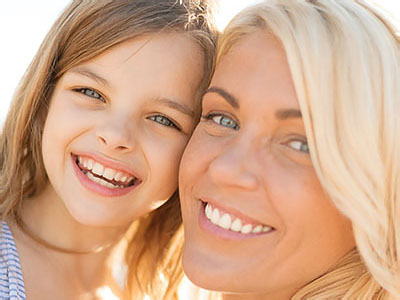 The image shows a woman with blond hair and a child standing close to her, both smiling at the camera. They are outdoors during daylight, as indicated by the bright lighting.