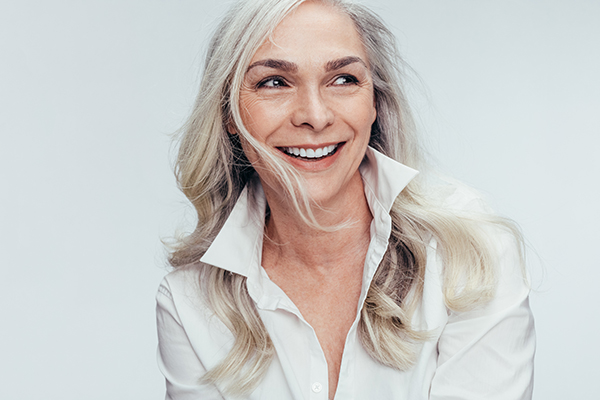 The image shows a woman with short hair smiling at the camera, wearing a white top and earrings, against a plain background.