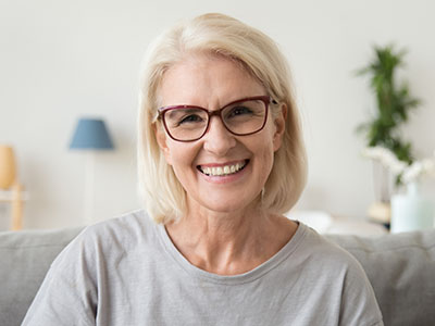 The image shows a woman with short blonde hair, wearing glasses and a light-colored top, smiling at the camera against a blurred indoor background.