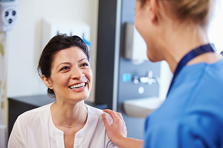 The image features two individuals in a medical setting  a woman with dark hair wearing a white top, smiling at another person who appears to be a nurse or healthcare professional, standing next to her with a stethoscope around their neck and a name tag on their shirt, both are engaged in a friendly interaction.