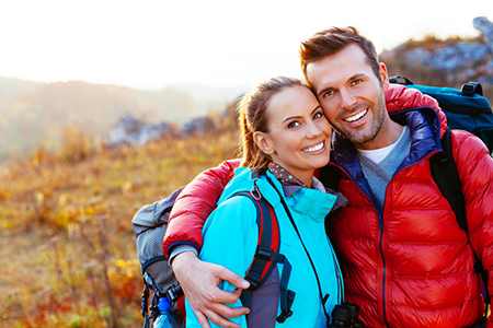A man and woman posing for a photo outdoors during daylight, smiling and embracing each other while wearing backpacks and hiking gear.