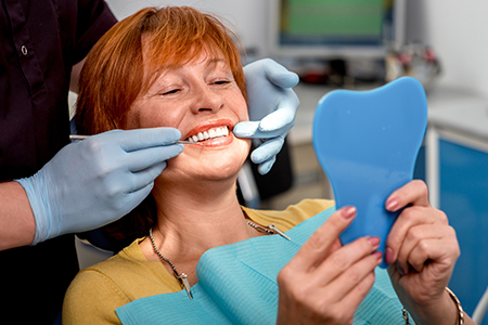 A woman sitting in a dental chair with a blue toothbrush-shaped object on her teeth, smiling at the camera while a dental professional works on her teeth.