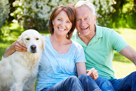 An elderly couple sitting on grass with a golden retriever dog between them, smiling and enjoying their time together outdoors.