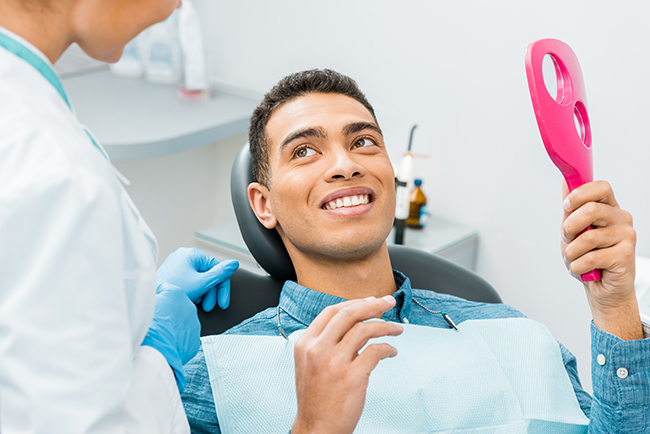The image shows a dental office setting with a smiling man sitting in the dentist s chair, receiving oral care from a dental professional who is using a pink instrument on his teeth.