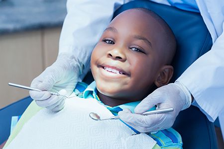 The image shows a young child sitting in a dental chair with a broad smile, receiving dental treatment from a dentist wearing gloves and a mask, who is seated behind the patient.