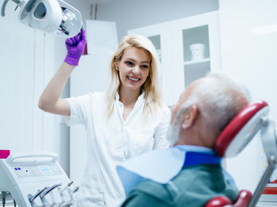 The image shows a woman standing next to an older man sitting in a dental chair, with both individuals wearing gloves  the woman appears to be a dentist or dental professional, and she is holding up a device that seems related to dental care.