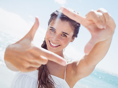 The image shows a young woman with long hair smiling at the camera while holding up two fingers, possibly for a  peace  sign, against a bright background that suggests a sunny beach setting.